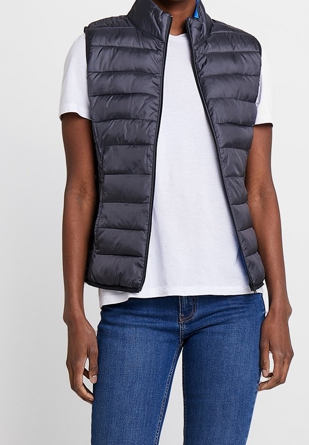 Personne portant une veste matelassée noire sans manches sur une chemise blanche à manches courtes et un jean bleu, debout, les mains détendues le long du corps.