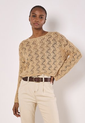 Knit sweater in light beige with an open lace pattern. Features long sleeves and a wide neckline, paired with high-waisted beige trousers and a brown belt.