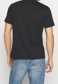 Muž v jednoduchém černém tričku s krátkým rukávem a vybledlých modrých džínsech, stojící s zády k divákovi na bílém pozadí.