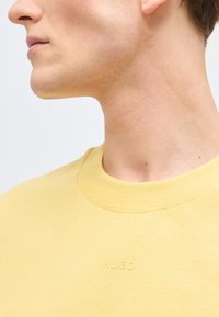 Νέος άνδρας με κίτρινο φούτερ με στρογγυλή λαιμόκοψη, με την επιγραφή "HUGO" ανάγλυφη στο στήθος, στρέφοντας ελαφρώς το κεφάλι στο πλάι, αποκαλύπτοντας τη γραμμή του σαγονιού και τον λαιμό.