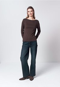 Jonge vrouw staat met handen achter haar rug, draagt een bruin langarmig shirt, donkerblauwe spijkerbroek en bruine enkellaarsjes.