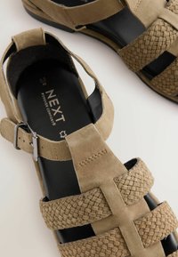 Sandale din suede bej cu bretele țesute, cataramă ajustabilă și o talpă interioară neagră. Prezintă laterale deschise și un design cu toc plat.