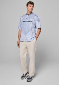 Jasnoniebieski jersey z krótkim rękawem w ukośne paski, czarny kołnierz i napis "WILLIAMS". Noszony z beżowymi spodniami oraz granatowo-szarymi sneakersami.