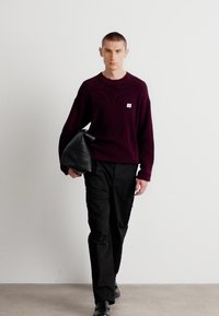 Maroon knitted sweater with a small logo, paired with black trousers and a black rectangular clutch bag, set against a light background.