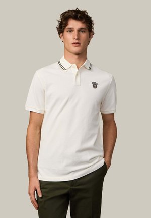 Witte polo met een zwart-wit gestreepte kraag, met een klein logo op de borst; korte mouwen en een zachte textuur.