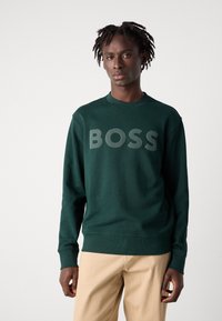 Felpa verde in tessuto morbido con scollo rotondo. Presenta un grande logo grigio "BOSS" sul davanti. Indossata con pantaloni beige.