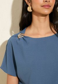 Top blu textured con design drappeggiato, caratterizzato da una spilla in accent oro sulla spalla. La modella indossa grandi orecchini in tono oro.