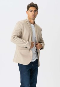 Beige blazer med strukturerede skuldre, enkeltknapslukning og to forlommer, parret med en hvid t-shirt og mørke jeans.
