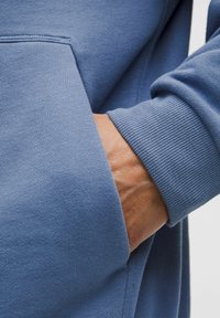 Jasnoniebieska bluza z gładkiego materiału, z kieszenią z przodu. W kieszeni spoczywa dłoń, widoczne są detale ściągacza.