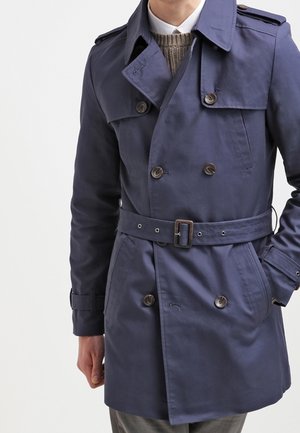 Hombre que lleva un abrigo trench azul marino doble botonadura con cinturón, un jersey marrón y una camisa blanca con cuello debajo.