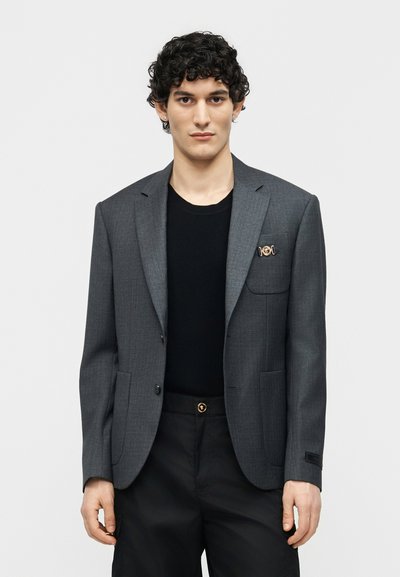 Jeune homme aux cheveux bouclés portant un blazer gris sur une chemise noire et un pantalon noir, debout devant un fond clair uni.
