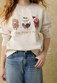 Sweatshirt crème avec trois motifs d'animaux festifs et texte doré "Joyeux Noël à Moo !" Orné de détails colorés. Jean bleu.