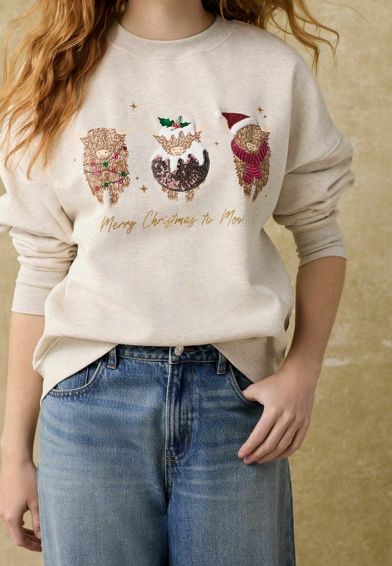 Sweatshirt crème avec trois motifs d'animaux festifs et texte doré "Joyeux Noël à Moo !" Orné de détails colorés. Jean bleu.