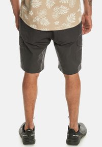 Black cargo shorts with multiple pockets, made from durable material; worn with a beige floral-patterned shirt and black shoes.