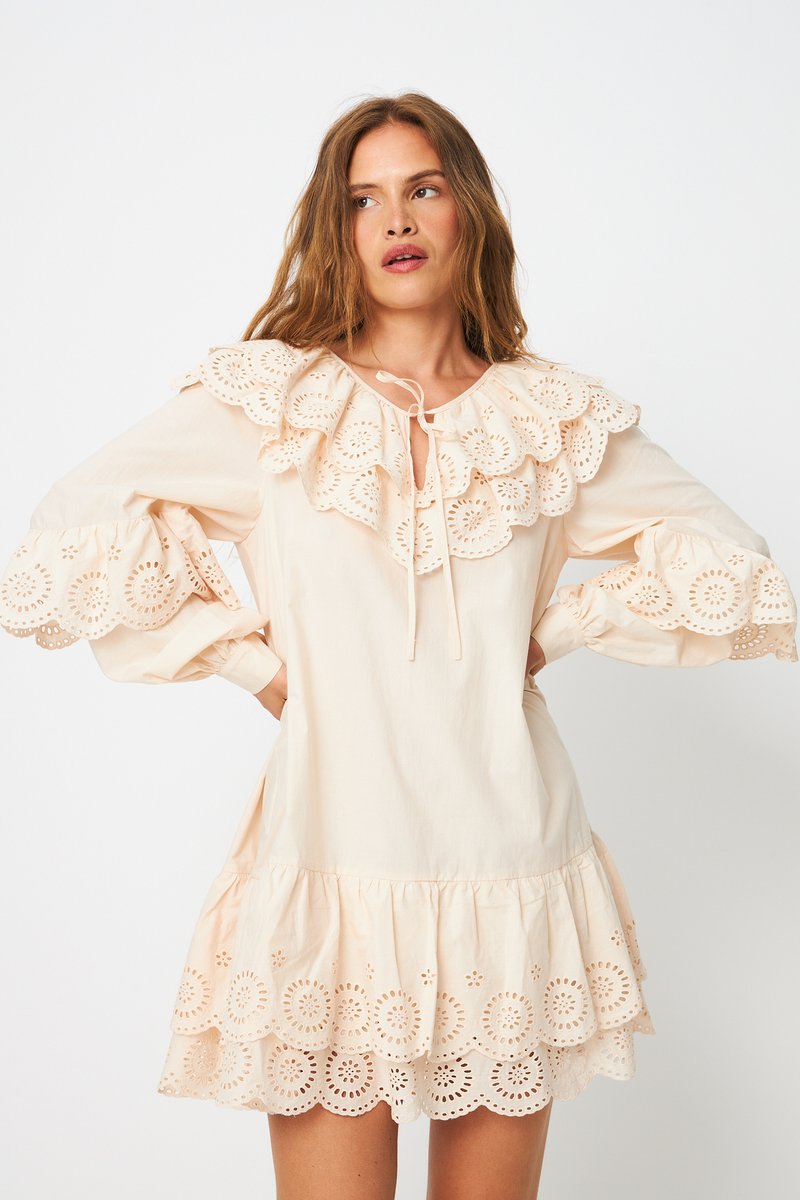 Γυναίκα με φόρεμα εκρού χρώματος με λεπτομέρειες από δαντέλα με τρύπες στον βολάν γιακά, τα μανίκια και το τελείωμα, στέκεται με τα χέρια στη μέση μπροστά σε λευκό φόντο.