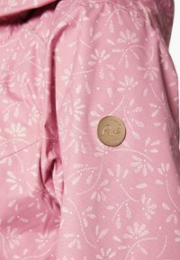 Ljuspink tygjacka med ett vitt blommönster, med en brun cirkulär logobadge på ärmen, som visar en slät textur.