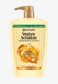 Bottiglia color miele con un tappo a pompa, caratterizzata da un motivo a nido d'ape e etichette nere e verdi, indicante shampoo per la riparazione dei capelli danneggiati.