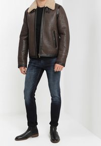 Brun läderbomberjacka med lammullskrage, dragkedjestängning och svarta läderdetaljer, kombinerad med mörka jeans och svarta stövlar.