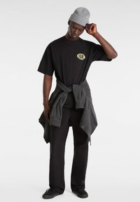 Camiseta negra de manga corta con el logo de "VANS", combinada con pantalones negros y una chaqueta de mezclilla gris atada, complementada con un gorro gris.