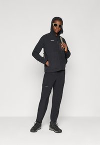 Giacca in pile nera con cappuccio e tasche con zip, abbinata a pantaloni neri con cerniere laterali e robuste scarpe da trekking nere.