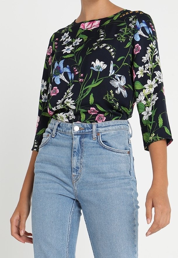 Femme portant un chemisier noir à fleurs à manches longues, rentré dans un jean bleu clair taille haute avec fermeture avant à boutons et zip.
