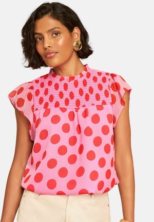 Vrouw met kort donker haar draagt grote gouden oorbellen en een roze blouse met rode stippen en gerimpelde korte mouwen.