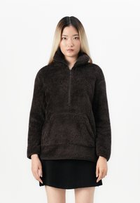 Brun fleecepullover med hög krage och kvartslång dragkedja. Mjuk, fluffig textur med rak nederkant och avslappnad passform. Kombineras med en svart kjol.