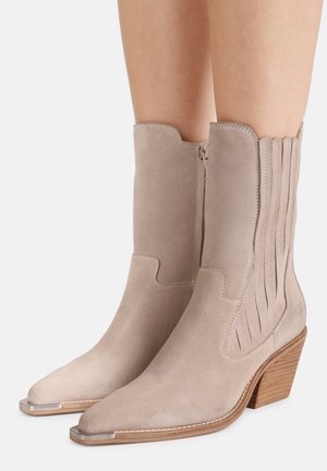 Beige suède laarzen tot halverwege de kuit met puntige metalen neuskapjes, ritssluiting aan de zijkant, decoratieve stiksels en gestapelde houten hakken, gedragen op blote benen.