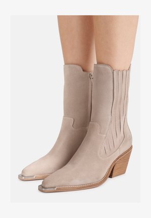 Beige suede mid-calf boots with pointed metal-toe caps, side zipper, decorative stitching, and stacked wooden heels worn on bare legs.