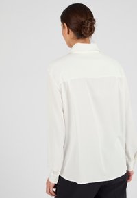 Blusa de manga larga, blanca, en tejido ligero con cuello tipo camisa, que presenta una textura suave y un dobladillo curvado en la parte trasera.