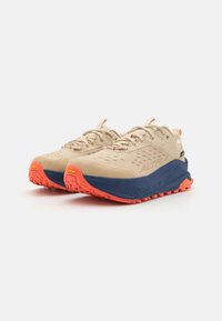 Beige joggesko med teksturert overdel, forsterket tå og særegen yttersåle. Har en navy mellomsole og lys oransje aksenter.