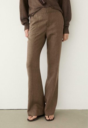 Donna che indossa pantaloni marroni a vita alta e gamba larga, abbinati a un maglione marrone lavorato a maglia, in piedi contro una parete bianca uniforme.