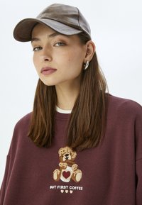 Gorra de cuero marrón, sudadera holgada color burdeos con un oso bordado sosteniendo un corazón y el texto: "PERO PRIMERO CAFÉ." Textura suave.