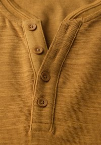 Detailní záběr na hořčičně žlutou texturovanou košili typu henley s čtyřmi hnědými knoflíky na žebrovém zapínání.