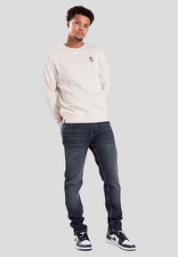 Light beige long-sleeve sweatshirt with small graphic on chest, paired with dark denim jeans and black and white high-top sneakers.