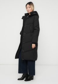 Kvinde iført en lang sort foret frakke med hætte, vide mørkeblå jeans og sorte hæl-støvletter, stående mod en ensfarvet hvid baggrund.