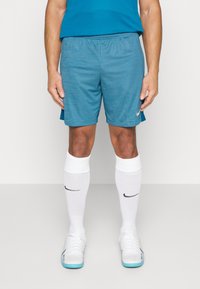 Hombre vistiendo pantalones cortos deportivos azules, calcetines Nike blancos hasta la rodilla y zapatillas deportivas blancas, de pie contra un fondo liso.