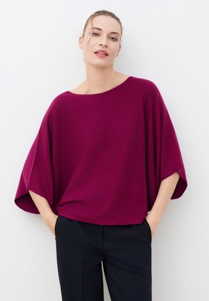Femme portant un haut ample magenta à larges manches et un pantalon noir, debout les mains dans les poches devant un fond blanc uni.
