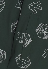 Zelená látka s opakujícím se stříbrným grafickým vzorem zakrytých postav a abstraktních tvarů. Na pravé straně je viditelná jedna kapsa.