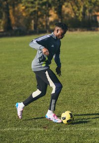 Urheilija harmaassa Adidas-harjoituspuvussa, jossa on fluoresoivat vihreät yksityiskohdat, mustissa housuissa, joissa on valkoiset raidat, ja värikkäissä jalkapallokengissä, jotka kuljettavat keltaista palloa.