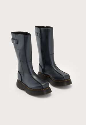 Bottes en cuir noires mi-mollet avec des semelles épaisses, coutures jaunes et fermetures éclair sur les côtés, sur un fond blanc uni.