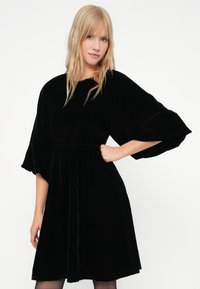 Rochie din catifea neagră cu mâneci scurte și largi, decolteu rotund și talie strânsă care se lărgește la tiv. Textură netedă.