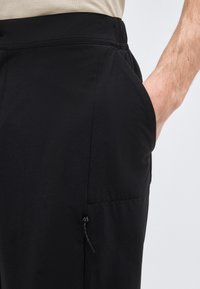 Gros plan sur une personne portant un pantalon noir avec une ceinture élastique et une poche latérale zippée, la main reposant à l'intérieur de la poche.