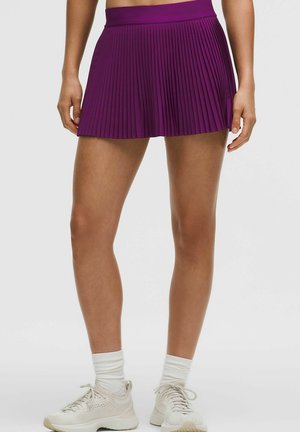 Persoană purtând adidași albi și șosete, asortați cu o fustă sportivă plisată mov, stând pe un fundal simplu.