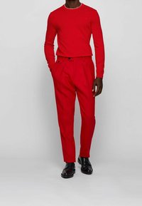 Maglia a maniche lunghe in maglia rossa abbinata a pantaloni rossi plissettati, con bottoni neri e scarpe eleganti nere, su sfondo neutro.