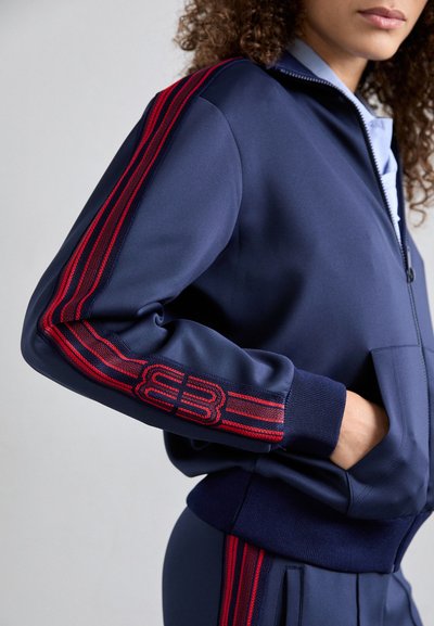 Giacca sportiva blu navy con accenti a strisce rosse sulle maniche, chiusura con zip e tasche laterali, realizzata in tessuto liscio ed elasticizzato.