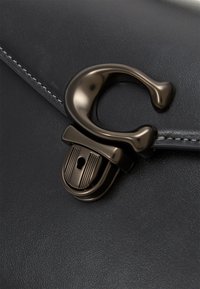 Musta nahkalaukku, jossa on näkyvä tummametallinen lukko avainaukko-yksityiskohdalla, sileä pinta ja reunassa kontrastompi ompelu.