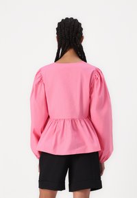 Blusa rosa con maniche a sbuffo, cintura aderente e orlo arricciato, realizzata in tessuto leggero. Indossata con pantaloni corti neri.