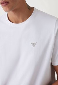 Hvid kortærmet t-shirt lavet af bomuld, med et lille gråt trekantet logo på brystet. Blød tekstur med rund halsudskæring.