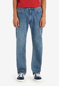 Personne portant un jean bleu ample, des baskets blanches et bleu marine, et un top rouge, debout les bras le long du corps sur un fond blanc.